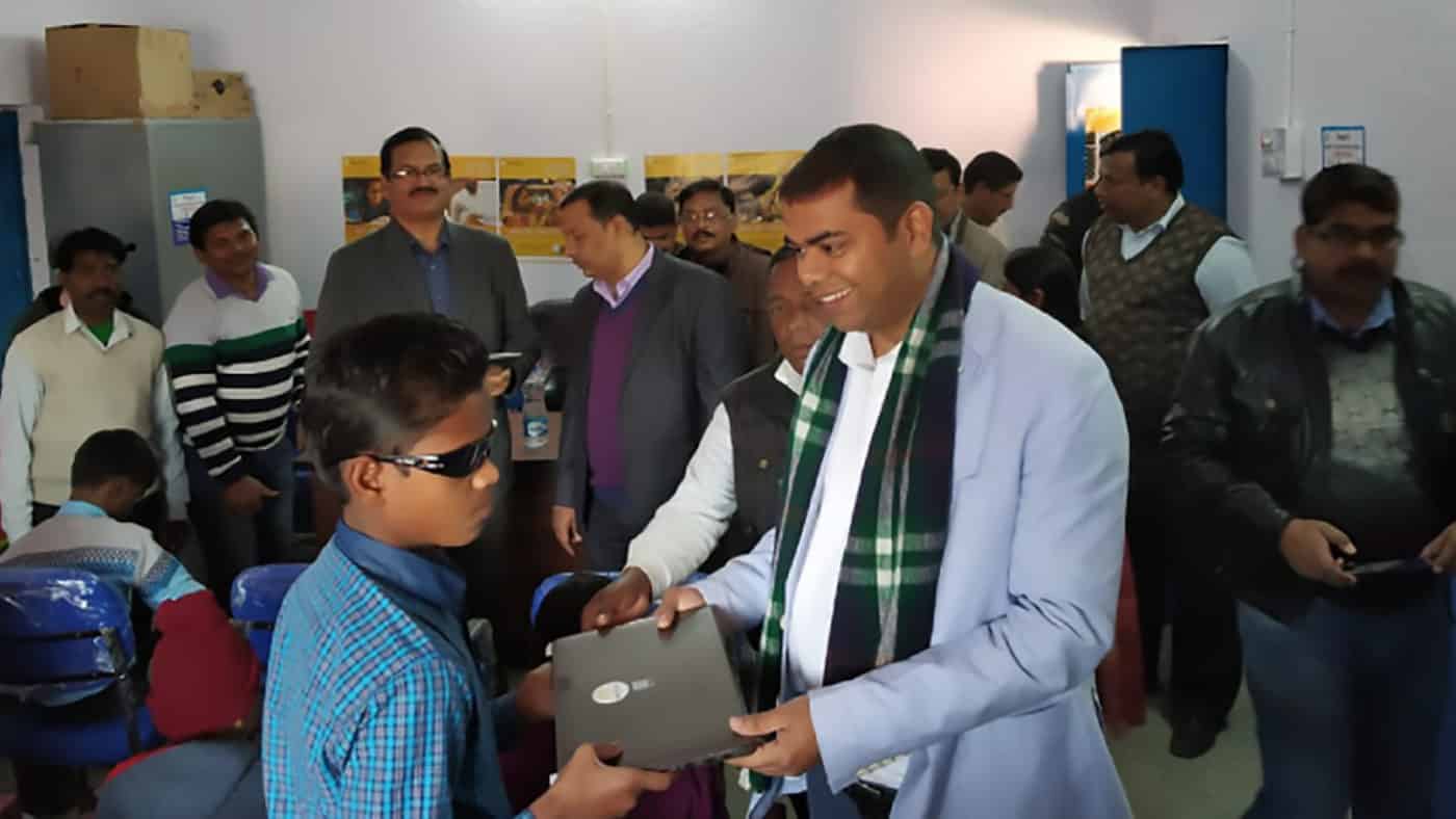 A man handing laptop to a boy with dark glasses on.