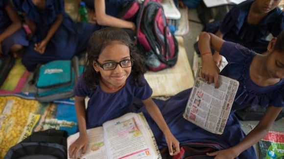 A girl sat on the floor with other school children around her, smiling.